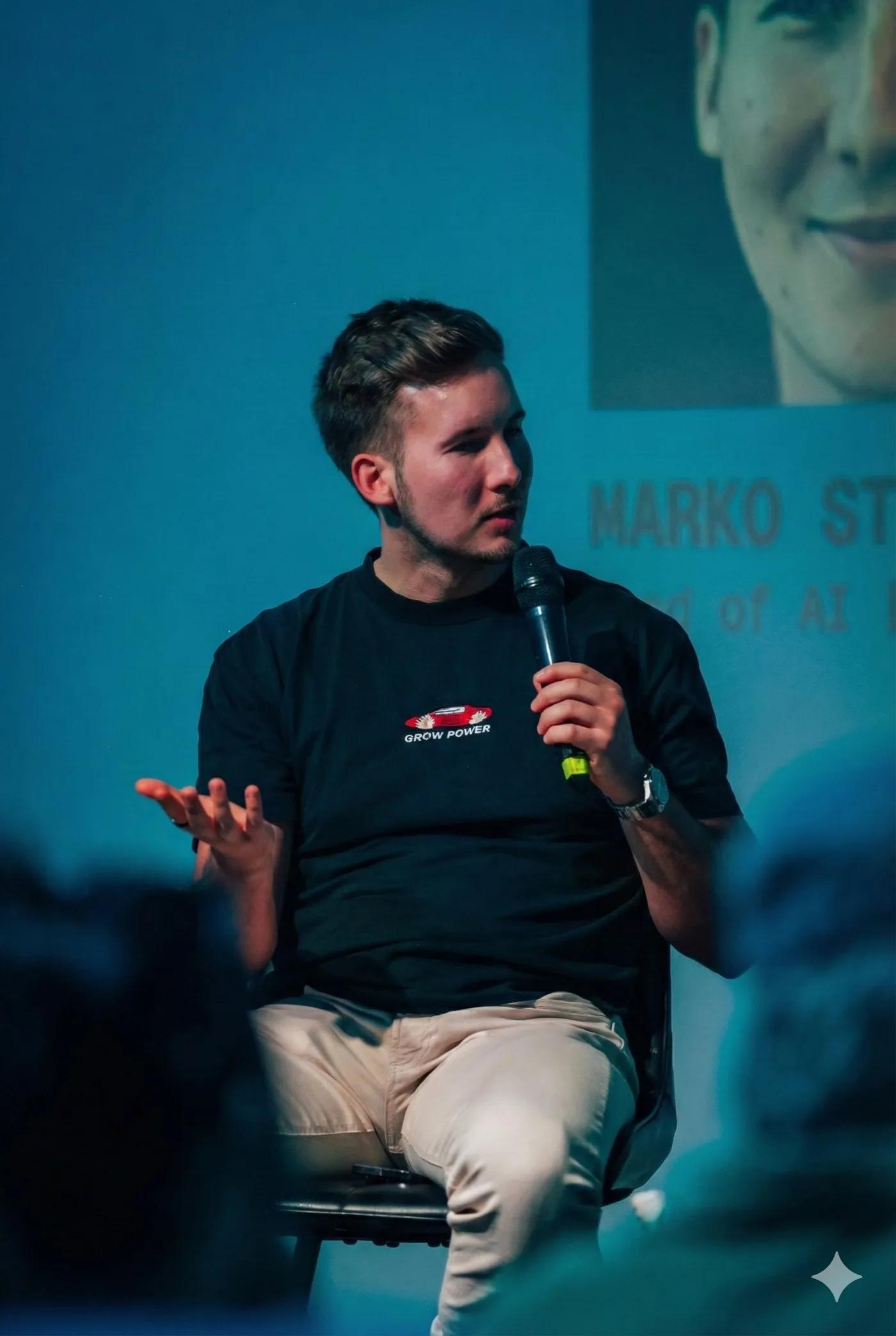 Marko on a stage chair with a microphone, explaining something with his hands, at the Open AGI Summit